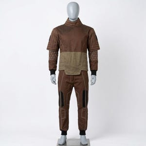 May include: A brown two-piece costume displayed on a mannequin. The top has a high collar and short sleeves with geometric stitching. The pants have black accents on the knees and ankles. The outfit is designed with a mix of brown and olive green panels.