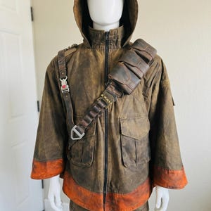 K&ouml;nnte beinhalten: Eine braune Kapuzenjacke mit Used-Optik, schwarzem Frontrei&szlig;verschluss, zwei gro&szlig;en Vordertaschen und einem breiten orangefarbenen Saum. &Uuml;ber der Brust befindet sich ein brauner, lederartiger Utility-Gurt mit Metallschnallen und Taschen.