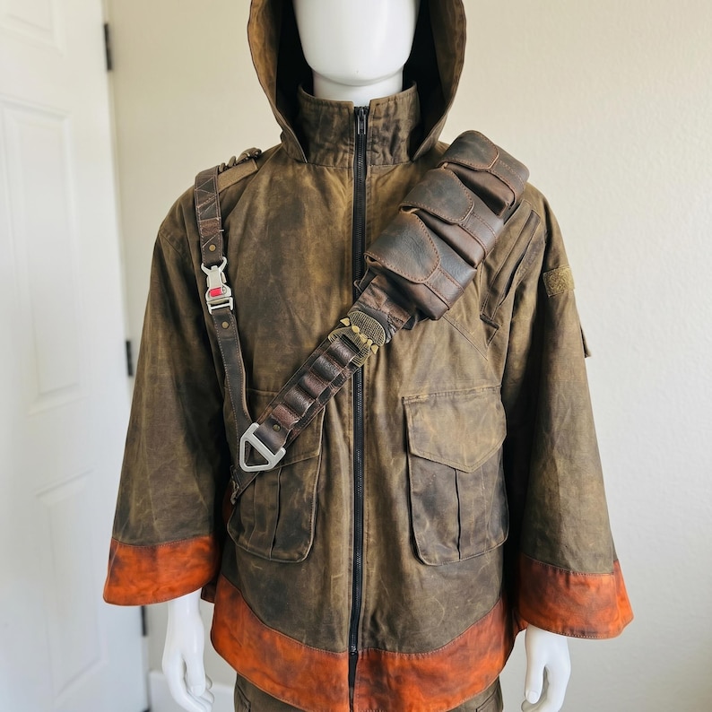 K&ouml;nnte beinhalten: Eine braune Kapuzenjacke mit Used-Optik, schwarzem Frontrei&szlig;verschluss, zwei gro&szlig;en Vordertaschen und einem breiten orangefarbenen Saum. &Uuml;ber der Brust befindet sich ein brauner, lederartiger Utility-Gurt mit Metallschnallen und Taschen.