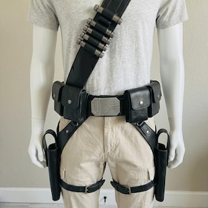 May include: A black utility belt system featuring a shoulder strap with silver cylindrical holders, a central buckle with geometric detailing, four rectangular pouches, and two drop-leg holsters secured with adjustable leg straps and buckles.
