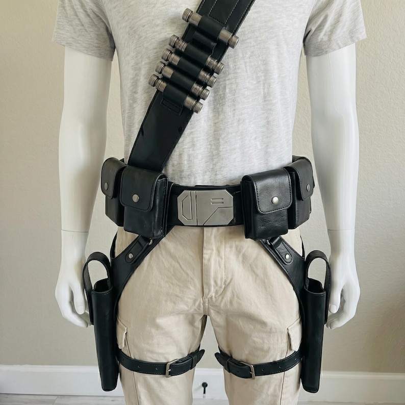May include: A black utility belt system featuring a shoulder strap with silver cylindrical holders, a central buckle with geometric detailing, four rectangular pouches, and two drop-leg holsters secured with adjustable leg straps and buckles.