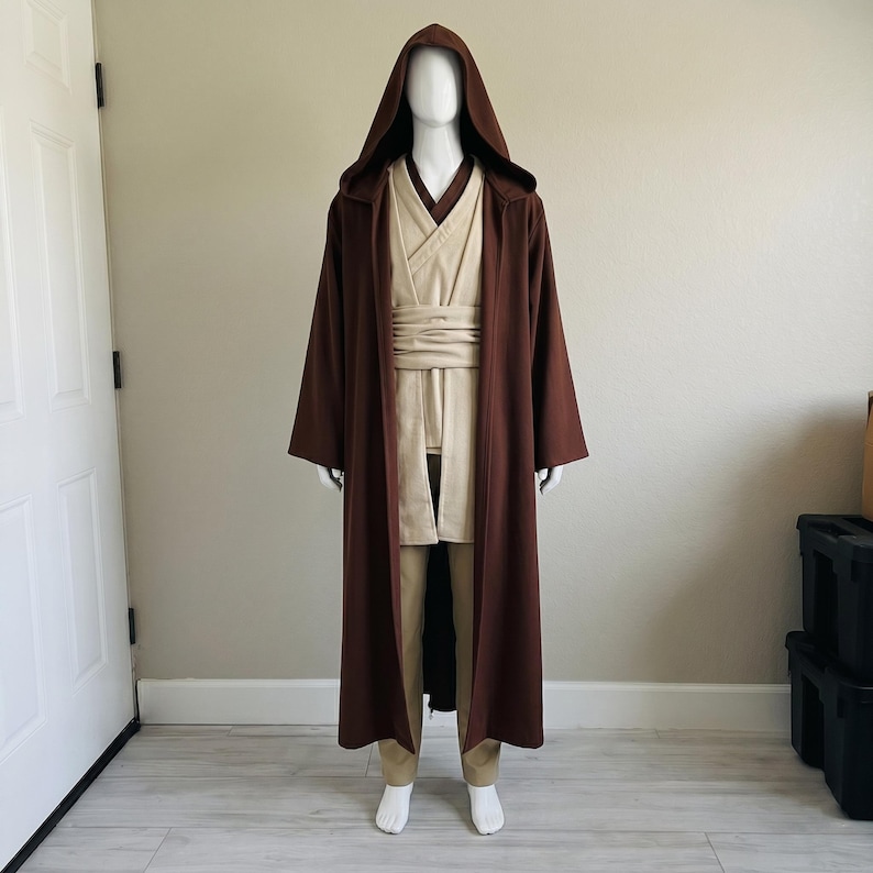 May include: A brown hooded robe costume with a beige tunic, trousers, and sash. The costume is displayed on a mannequin. The robe is long and flowing, with wide sleeves. The tunic has a V-neck and the trousers are a neutral colour.