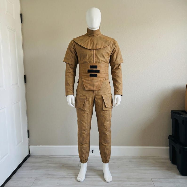 May include: A tan costume on a mannequin. The outfit features a high-collared top with short sleeves, a wide belt with black details, and matching trousers with pockets. The costume is made of a textured fabric.