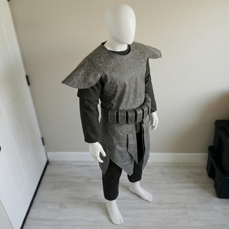 May include: A gray costume with shoulder pads, a tunic, and a belt. The costume is displayed on a mannequin. The outfit includes black pants and a long-sleeved shirt. The costume is made of a textured material.