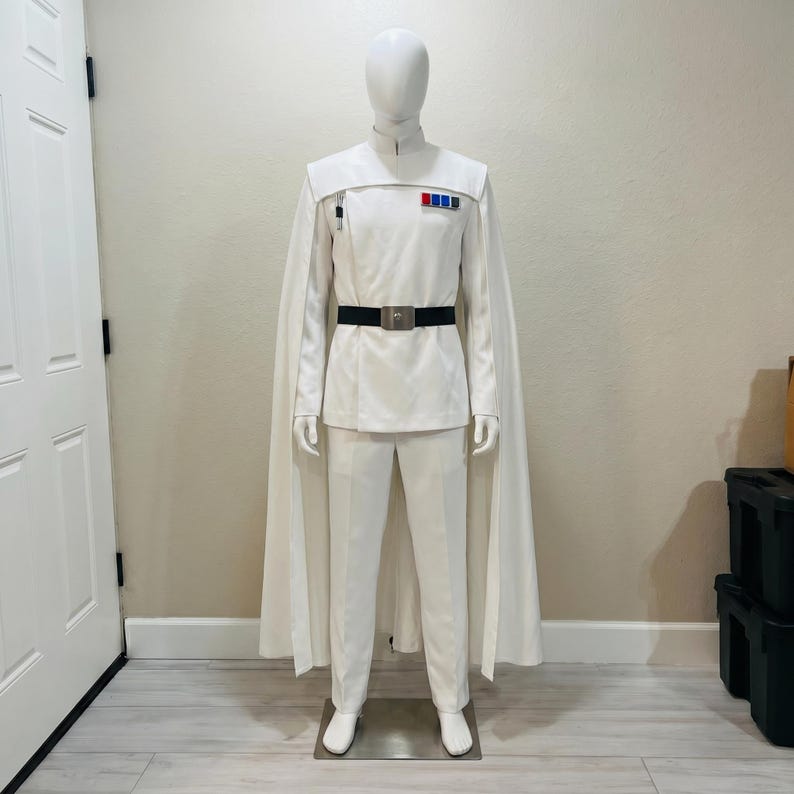 May include: A white costume with a high collar, cape, and black belt. The outfit includes a jacket with a small pocket and a row of coloured pins. The trousers are white, and the costume is displayed on a mannequin.