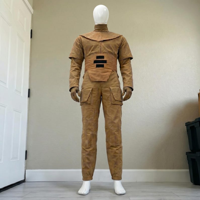 K&ouml;nnte beinhalten: Ein hellbraunes Kost&uuml;m, das auf einer Schaufensterpuppe pr&auml;sentiert wird. Das Outfit umfasst ein Oberteil mit hohem Kragen und breitem Schulterteil, einen G&uuml;rtel mit zwei horizontalen schwarzen Streifen und eine passende Hose mit Taschen. Die Schaufensterpuppe tr&auml;gt auch Handschuhe.