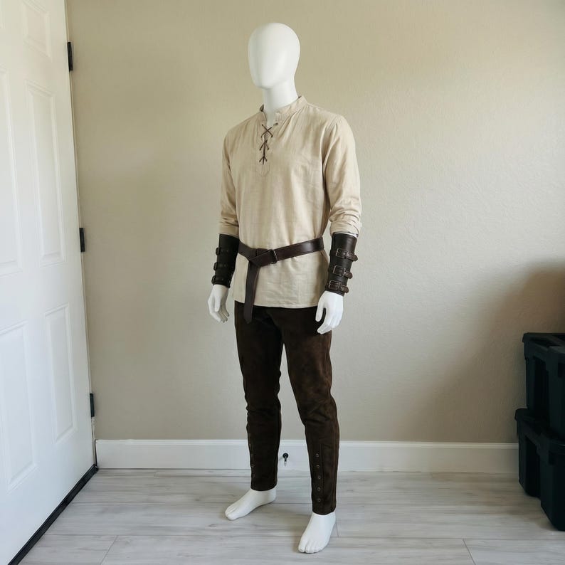Puede incluir: Una camisa beige de manga larga con cuello acordonado, pantalones de color marr&oacute;n oscuro, un cintur&oacute;n de cuero marr&oacute;n oscuro y brazaletes de antebrazo de cuero marr&oacute;n oscuro a juego con hebillas, sobre un maniqu&iacute; blanco.