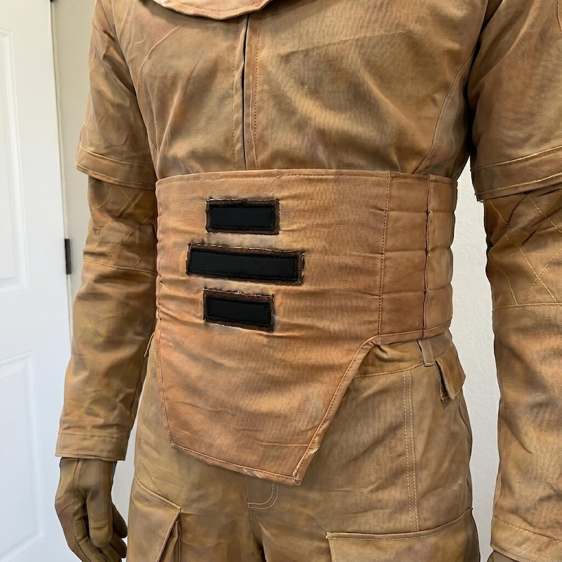 K&ouml;nnte beinhalten: Ein beigefarbenes Kost&uuml;m mit einem gepolsterten G&uuml;rtel mit drei schwarzen rechteckigen Akzenten. Das Outfit umfasst ein Oberteil mit Kragen, lange &Auml;rmel und eine Hose mit Taschen. Die Handschuhe sind beige.