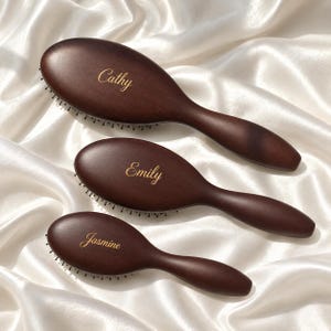 May include: Three dark brown wooden hairbrushes of varying sizes, each featuring a smooth, rounded handle and head. The brushes are engraved with the names Cathy, Emily, and Jasmine in a gold-colored script font on the back of the brush head.