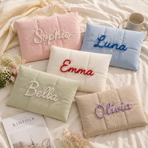 Pode incluir: Cinco almofadas retangulares acolchoadas em tons pastel, cada uma com um nome bordado em letras cursivas em relevo. As almofadas são rosa com Sophie, azul com Luna, branca com Emma, verde-sálvia com Bella e bege com Olivia.