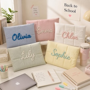Pode incluir: Cinco estojos retangulares acolchoados em azul claro, rosa, branco, verde sálvia e amarelo pálido. Cada estojo apresenta um nome em letras cursivas em relevo: Olivia, Emma, Chloe, Lily e Sophie. Os estojos estão dispostos sobre uma mesa.