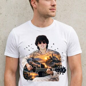 May include: A white short-sleeved t-shirt featuring a graphic print of a person with dark hair and sunglasses playing an acoustic guitar. The guitar body and the person's torso are filled with a scenic landscape illustration of a castle at sunset.