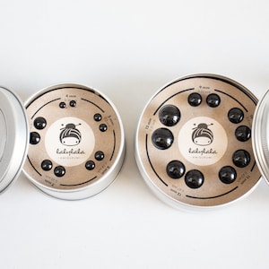 Puede incluir: Tres contenedores redondos con botones negros de diferentes tamaños. Cada contenedor tiene una etiqueta con el tamaño de los botones en milímetros y pulgadas. Las etiquetas también incluyen el texto "lalylala AMIGURUMI".