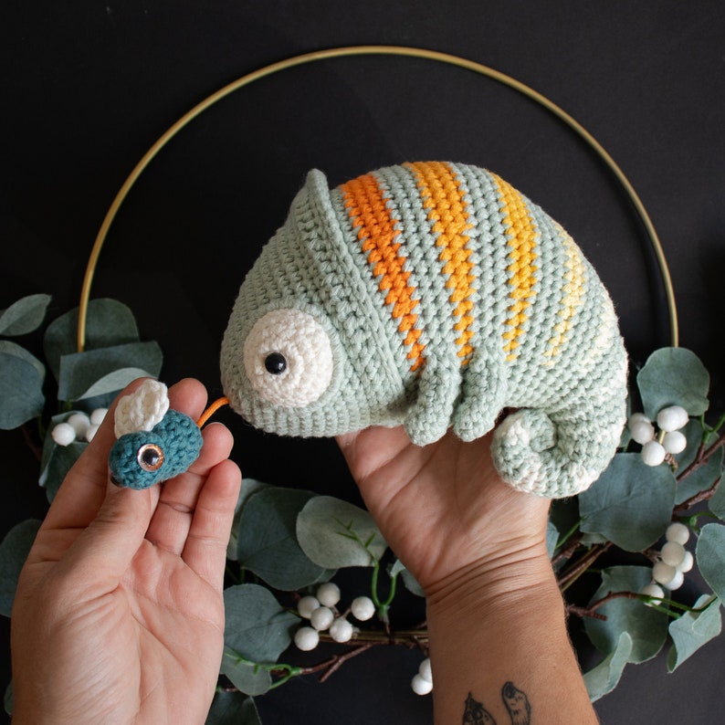Puede incluir: Un camale&oacute;n de crochet verde y naranja con una mosca de crochet azul y verde. El camale&oacute;n tiene un ojo blanco y est&aacute; sentado en una mano.