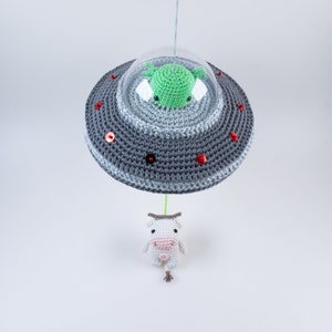 May include: A crocheted gray UFO with a green alien inside and red sequins. The UFO is hanging from a string above a white crocheted cow.