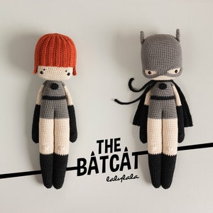 May include: Two crocheted stuffed toys, one with a red head and the other with a gray cape, both wearing black outfits. The text "THE BATCAT babylala" is below the toys.