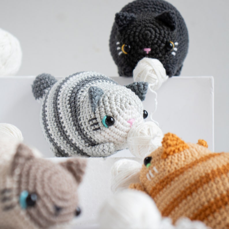 Puede incluir: Cuatro juguetes de crochet con forma de gato, cada uno con un color y un patr&oacute;n diferente. Los gatos sostienen ovillos de lana. Los gatos est&aacute;n sentados sobre una superficie blanca.