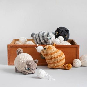 Puede incluir: Cinco juguetes de crochet con forma de gato en varios colores, incluyendo gris, negro, naranja y beige, est&aacute;n dispuestos en una caja de madera con bolas de lana blanca. Los gatos est&aacute;n todos sentados y mirando a la c&aacute;mara.