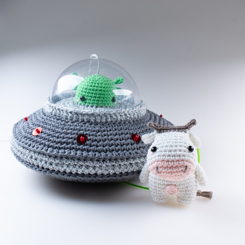 May include: A gray crocheted UFO with red beads and a green crocheted alien inside. A white crocheted cow with pink accents is standing in front of the UFO.