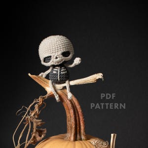 Puede incluir: Una pequeña figura de esqueleto tejida a ganchillo con un cráneo beige y un torso negro con detalles de costillas en blanco. La figura está sentada sobre el tallo de una calabaza. El texto PDF PATTERN es visible a la derecha sobre fondo oscuro.
