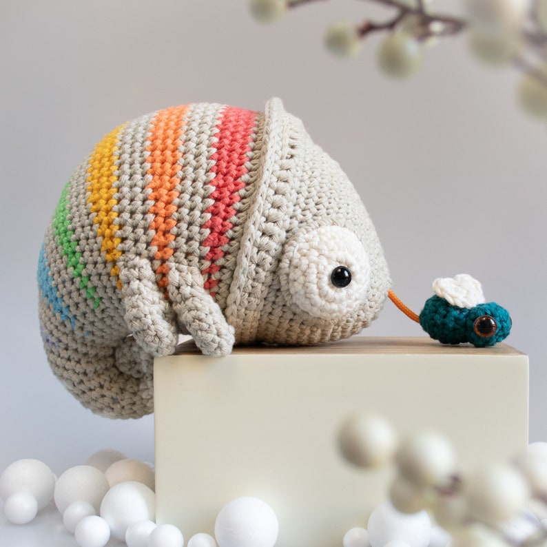 Puede incluir: Un camale&oacute;n de crochet con rayas arco&iacute;ris, sentado en una superficie blanca, mirando a una peque&ntilde;a mosca verde de crochet con ojos negros.