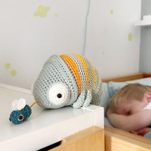 Puede incluir: Un juguete de camale&oacute;n de crochet con rayas naranjas y verdes, sentado sobre una superficie blanca. Un peque&ntilde;o juguete de mosca azul de crochet est&aacute; en primer plano.