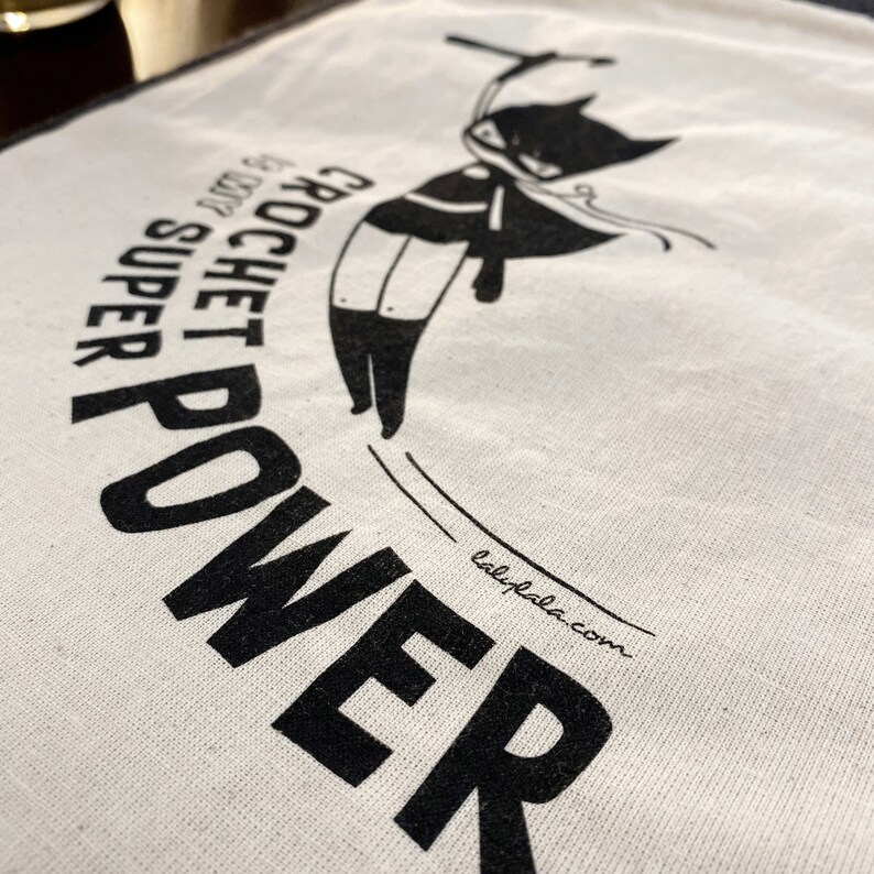 Puede incluir: Una bolsa de tela blanca con una ilustraci&oacute;n negra de un gato con una capa y una m&aacute;scara. El texto "CROCHET SUPER POWER" est&aacute; impreso en negro en la bolsa. El sitio web "lalypala.com" est&aacute; impreso en negro en la parte inferior de la bolsa.