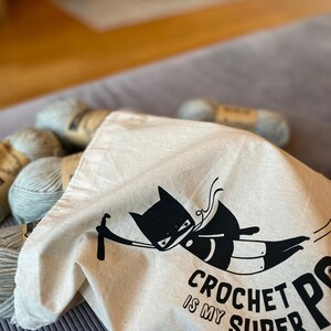Puede incluir: Una bolsa de lona blanca con un dise&ntilde;o de superh&eacute;roe de gato negro y el texto "Crochet is my super power". La bolsa est&aacute; llena de ovillos de lana.