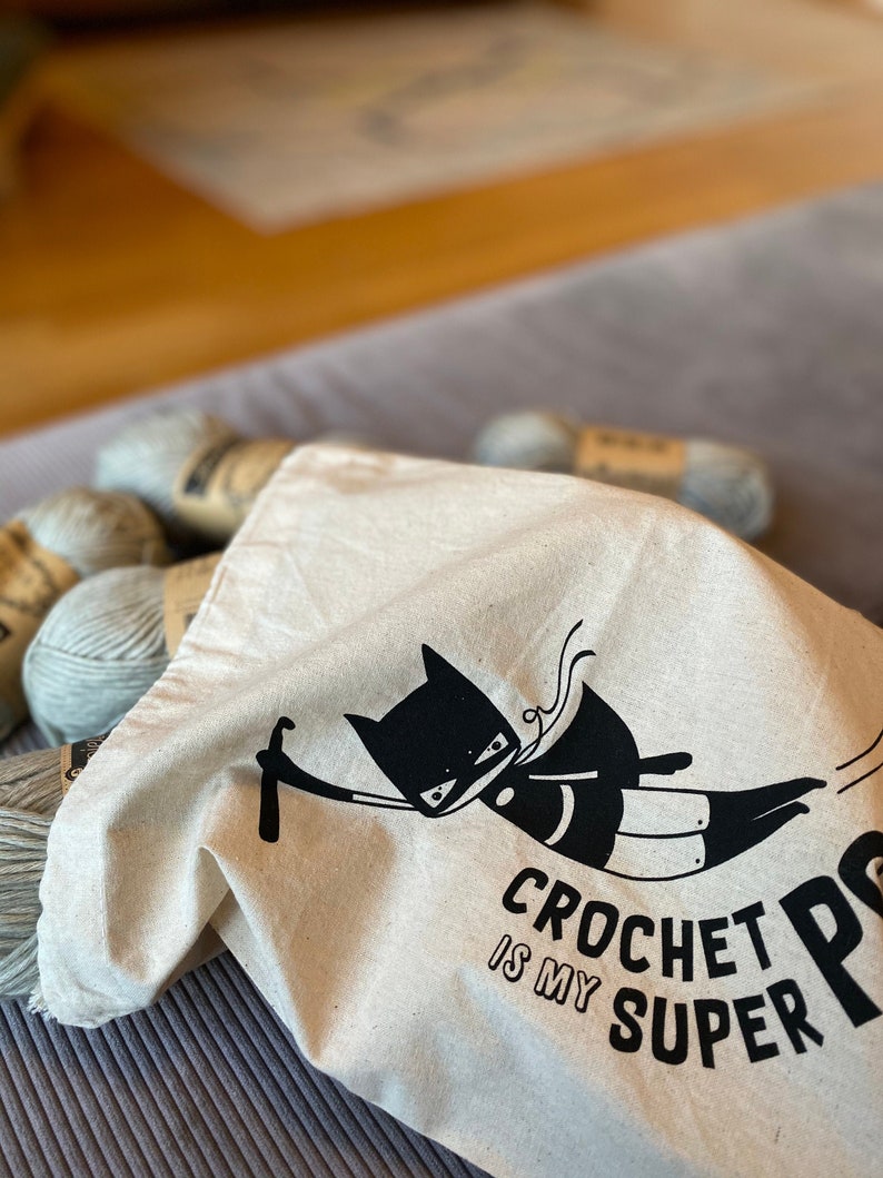 Puede incluir: Una bolsa de lona blanca con un dise&ntilde;o de superh&eacute;roe de gato negro y el texto "Crochet is my super power". La bolsa est&aacute; llena de ovillos de lana.