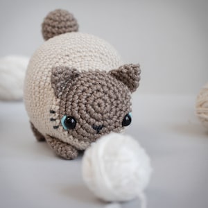 Puede incluir: Un juguete de gato de crochet con pelo marr&oacute;n y beige, ojos azules y una bola de lana blanca.