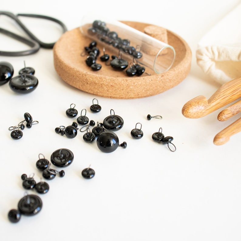 May include: A close-up of a glass jar filled with black buttons with small metal loops. The buttons are scattered on a cork coaster and a white surface.