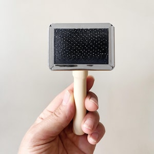 Puede incluir: Un cepillo carda rectangular para mascotas con un cabezal de metal plateado, cerdas negras finas y densas, y un mango de madera clara y lisa. El cabezal del cepillo tiene esquinas redondeadas y un acabado pulido para el aseo.