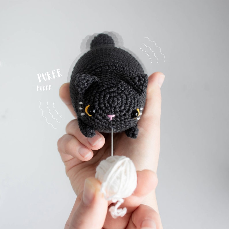 Puede incluir: Un juguete de gato de crochet negro con ojos amarillos y una nariz rosa. El gato est&aacute; sosteniendo una bola de hilo blanco en sus patas. El texto "PURRR" y "PURRR" est&aacute; escrito en blanco sobre un fondo gris.