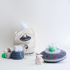May include: A crocheted UFO toy with a green alien inside, a crocheted cow toy, and a white canvas bag with a black and white UFO graphic and the text "Space for Unfinished Objects".