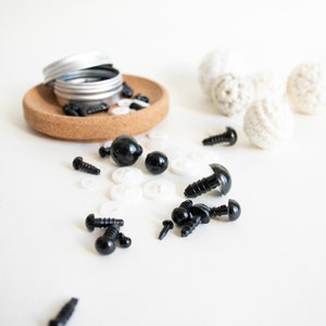 May include: A collection of black plastic safety eyes for crafts, with a variety of sizes and styles, including some with washers. The eyes are scattered on a white surface, along with some white plastic washers.