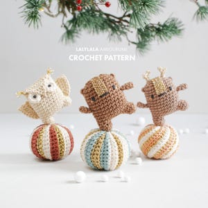 Op de afbeelding: Drie gehaakte amigurumi-speelgoedjes, een uil, een beer en een rendier, die elk op een gestreepte bal staan. De ballen zijn gehaakt in beige, bruin en gouden tinten.