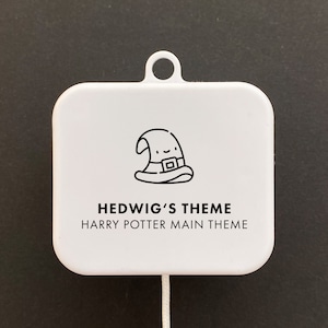 Könnte beinhalten: Weiße quadratische Musikbox mit einer Zugschnur. Die Box hat eine schwarze Strichzeichnung eines Zaubererhutes mit einem Gesicht. Der Text "Hedwig's Theme Harry Potter Main Theme" ist unter der Zeichnung gedruckt.