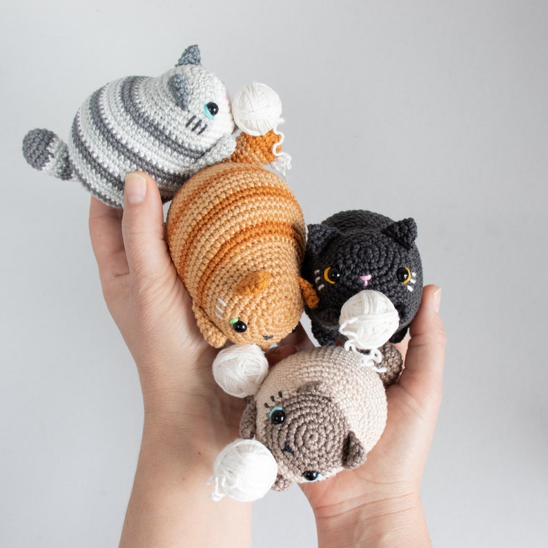 Puede incluir: Cuatro juguetes de gato de crochet con ovillos de lana. Los gatos son grises y blancos, naranjas y blancos, negros y marrones y blancos.