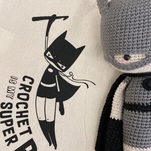Puede incluir: Una bolsa de lona beige con una ilustraci&oacute;n en blanco y negro de un superh&eacute;roe colgado de un gancho con el texto "CROCHET IS MY SUPER POWER". Un mu&ntilde;eco de Batman de crochet gris est&aacute; en el fondo.
