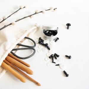 May include: A set of black plastic safety eyes, white plastic washers, and a small tin container.  The eyes and washers are scattered on a white surface.  There are also a pair of scissors, a white fabric bag, and four wooden crochet hooks.