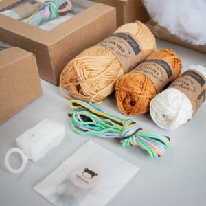 Puede incluir: Un kit de manualidades con tres ovillos de lana en beige, naranja y blanco. La lana est&aacute; etiquetada como "Catona" y "Simply Soft". El kit tambi&eacute;n incluye una peque&ntilde;a bolsa de hilo de bordar de colores, un anillo de pl&aacute;stico blanco y una pieza de pl&aacute;stico blanca con dos agujeros.