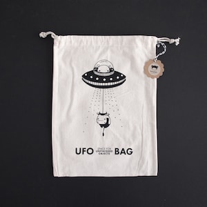 May include: White cotton drawstring bag with a black and white illustration of a UFO abducting a cow. The bag has the text "UFO BAG" and "SPACE FOR UNFINISHED OBJECTS".