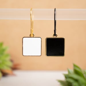 May include: A pair of square earrings, one white and one black, with gold-colored frames and ear wires. The earrings are hanging on a white rod against a blurred, neutral background. The earrings are a simple, modern design.