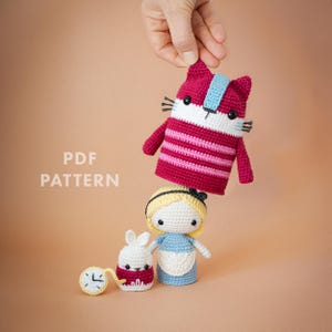 以下が含まれることがあります： 不思議の国のアリスのキャラクターの編み人形。 ピンクと白の縞模様のチェシャ猫、青いドレスのアリス、白いウサギ、懐中時計が含まれています。 左側には「PDF PATTERN」の文字が印刷されています。