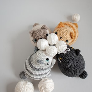 Puede incluir: Cuatro juguetes de crochet con forma de gato en varios colores, incluyendo gris, marr&oacute;n, naranja y negro, con ovillos de lana blanca esparcidos a su alrededor.