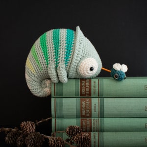 Puede incluir: Un juguete de camale&oacute;n de crochet verde y azul sentado encima de una pila de libros verdes con el texto "Brehms Tierleben" en los lomos. Una peque&ntilde;a mosca de crochet azul y verde est&aacute; sentada en el libro superior.