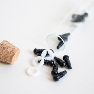 May include: A glass test tube filled with black plastic safety eyes and white plastic washers. The test tube is tipped over and the eyes and washers are spilling out onto a white surface.