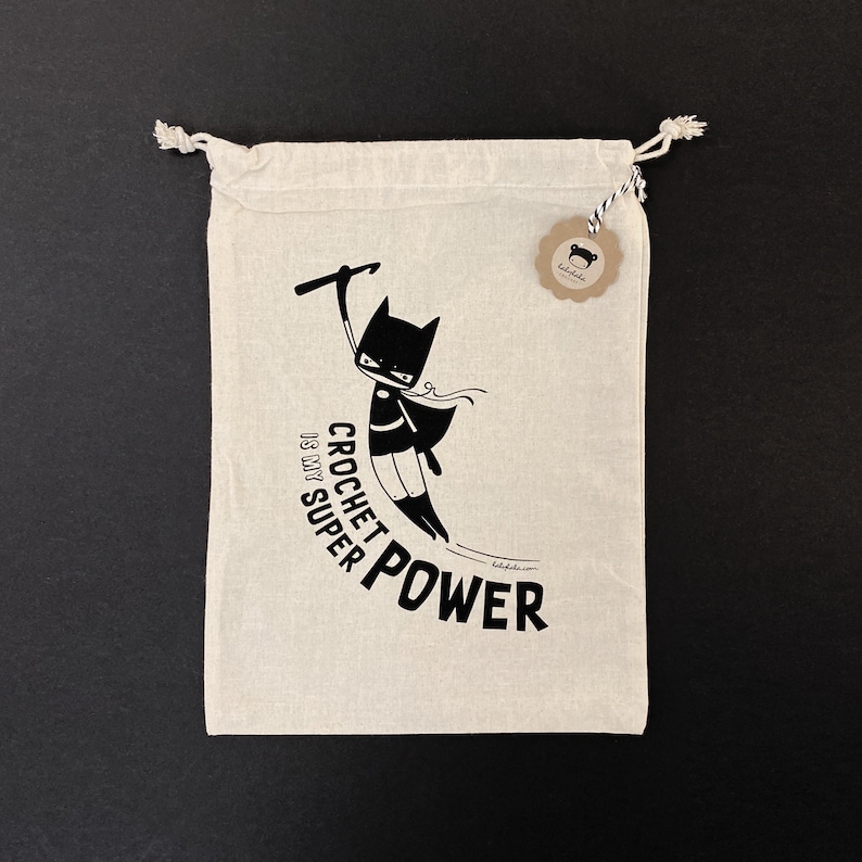Puede incluir: Una bolsa de algod&oacute;n blanco con cord&oacute;n de ajuste, ilustrada en blanco y negro con un superh&eacute;roe que sostiene un gancho de ganchillo. El texto "Crochet is my super power" est&aacute; impreso en la bolsa.