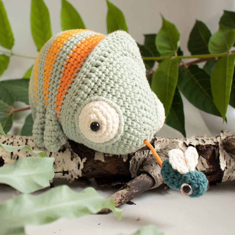Puede incluir: Un camale&oacute;n verde de crochet con rayas naranjas en la espalda est&aacute; posado en una rama, con la lengua extendida para atrapar una mosca verde de crochet.