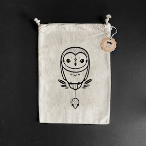 Könnte beinhalten: Eine weiße Baumwolltasche mit Kordelzug, die mit einer schwarz-weißen Illustration eines Eulen, die eine Maus hält, verziert ist. Die Tasche hat ein braunes Etikett mit dem Text "tefaloka" darauf.